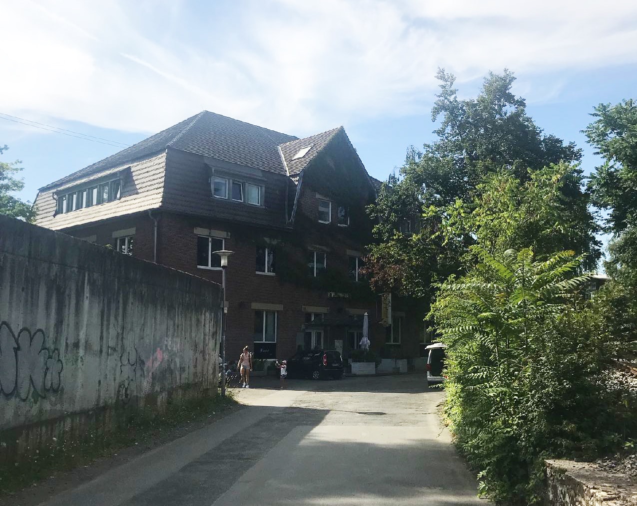 Zufahrt zum Büro: links Betonwand, rechts Bäume, am Ende dreigeschossiger Bau, Zugang frontal