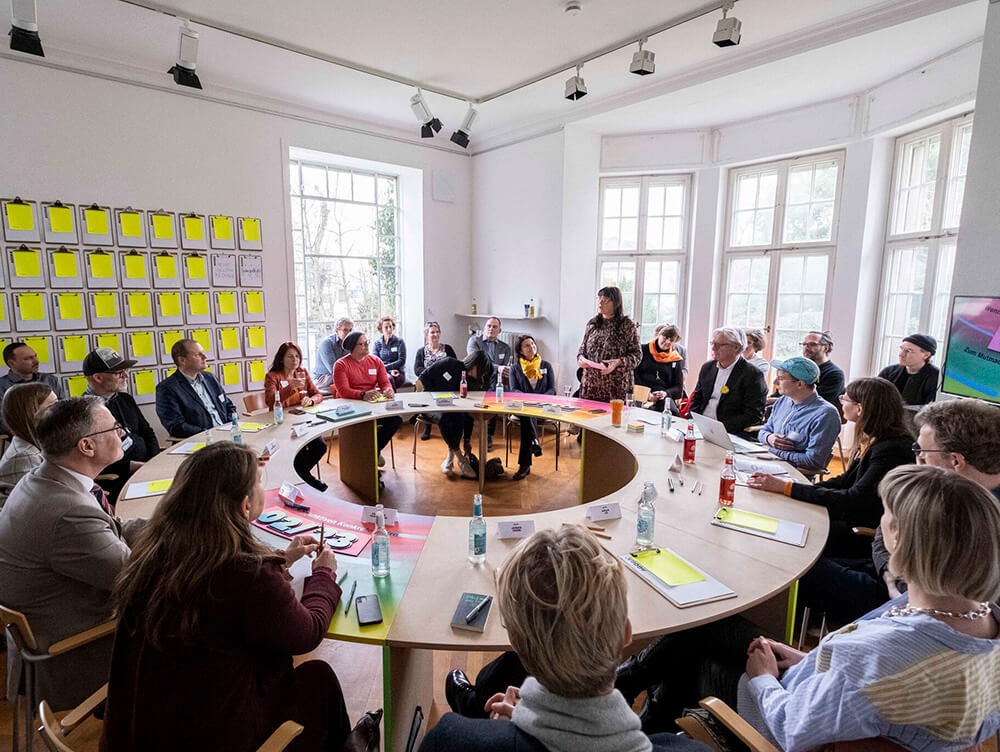 Menschen sind um einen kreisrunden Tisch versammelt, eine Person steht und spricht. Dahinter, in zweiter Reihe sitzen weitere Menschen auf Stühlen. 
