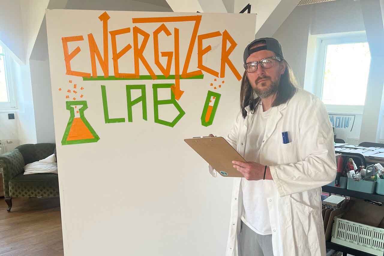 Mann mit Brille, langem Haar und weißem Laborkittel hält Klemmbrett vor Wand mit Schriftzug 'Energizer Lab' und Zeichnungen von Reagenzglas und Batterie