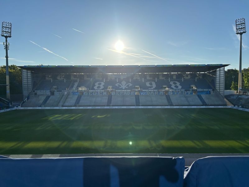 Leeres Fußballstadion mit Tribüne, auf der die Jahreszahlen 1898 und ein Lilien-Logo zu sehen sind, bei Sonnenaufgang