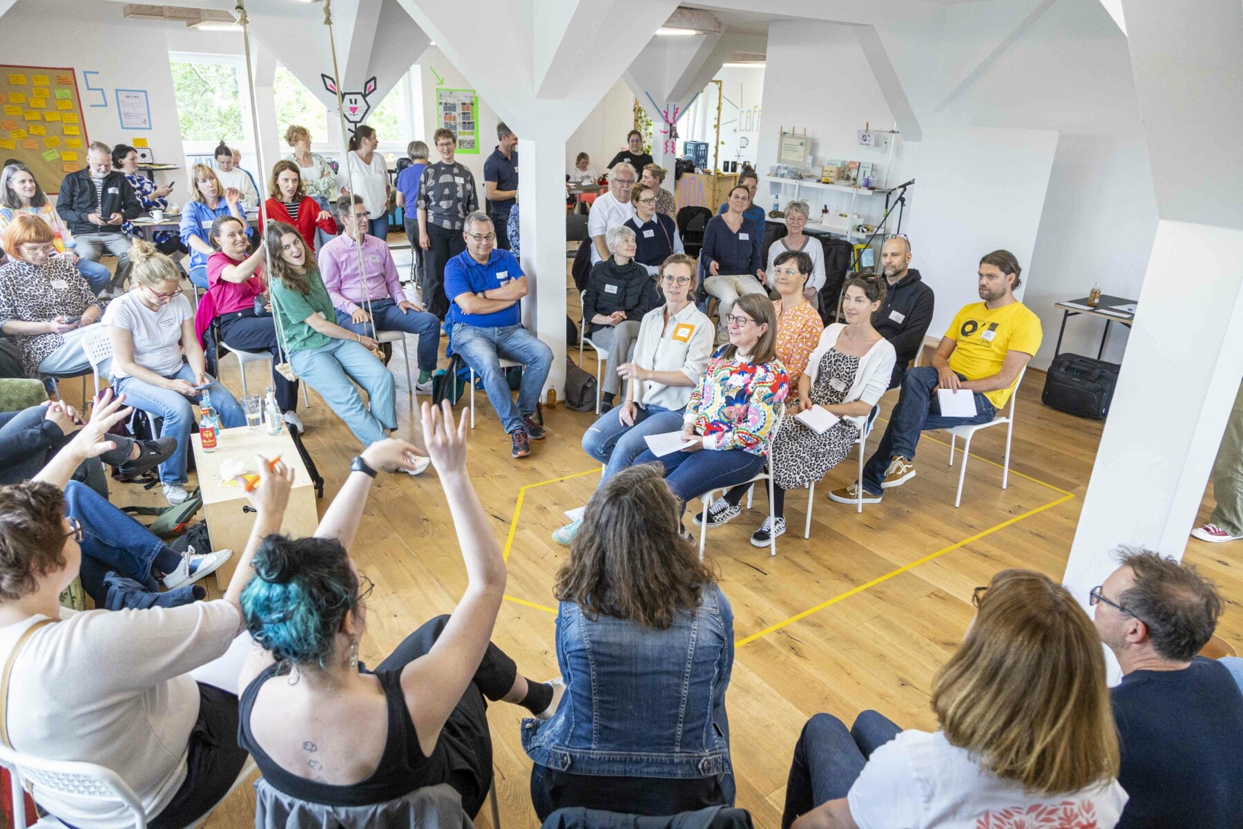 Teilnehmende eines Workshops sitzen in einem großen Raum in einem Stuhlkreis, in der Mitte des Kreises ist eine Stuhlreihe mit 6 Stühlen und Menschen aufgebaut, einige sprechen und andere hören aufmerksam zu