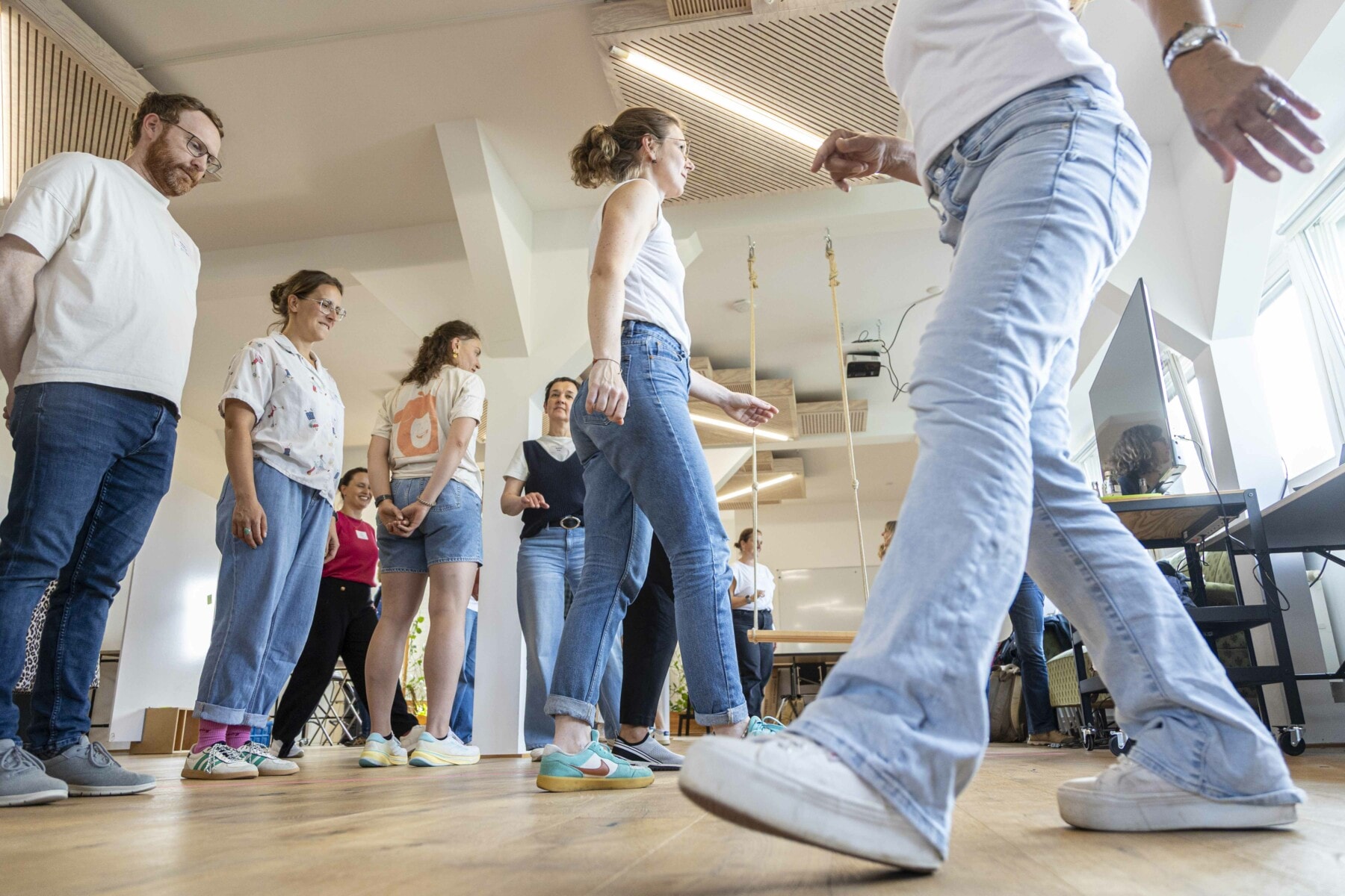 Teilnehmende eines Bewegungs-Workshops auf der KOON25 stehen in lockerer Formation in einem hellen Raum mit Holzboden und weißen Wänden. Einige laufen auch im Raum umher, sodass im Vordergrund ein Bein mit Sneaker-Schuh gezeigt ist.