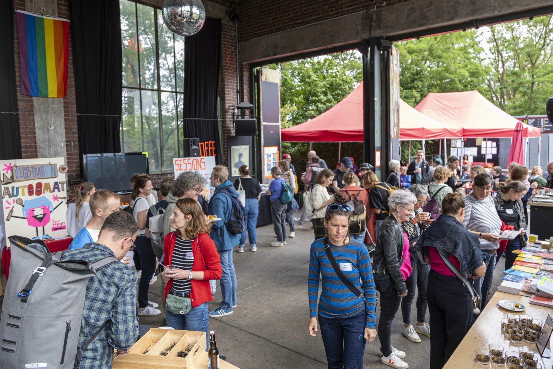 Das Bild zeigt die Halle der Community Area der KOON25 mit vielen Menschen, die an Ständen mit Büchern und Illustrationen stöbern oder sich in Kleingruppen unterhalten. Im Hintergrund sind rote Zelte zu sehen