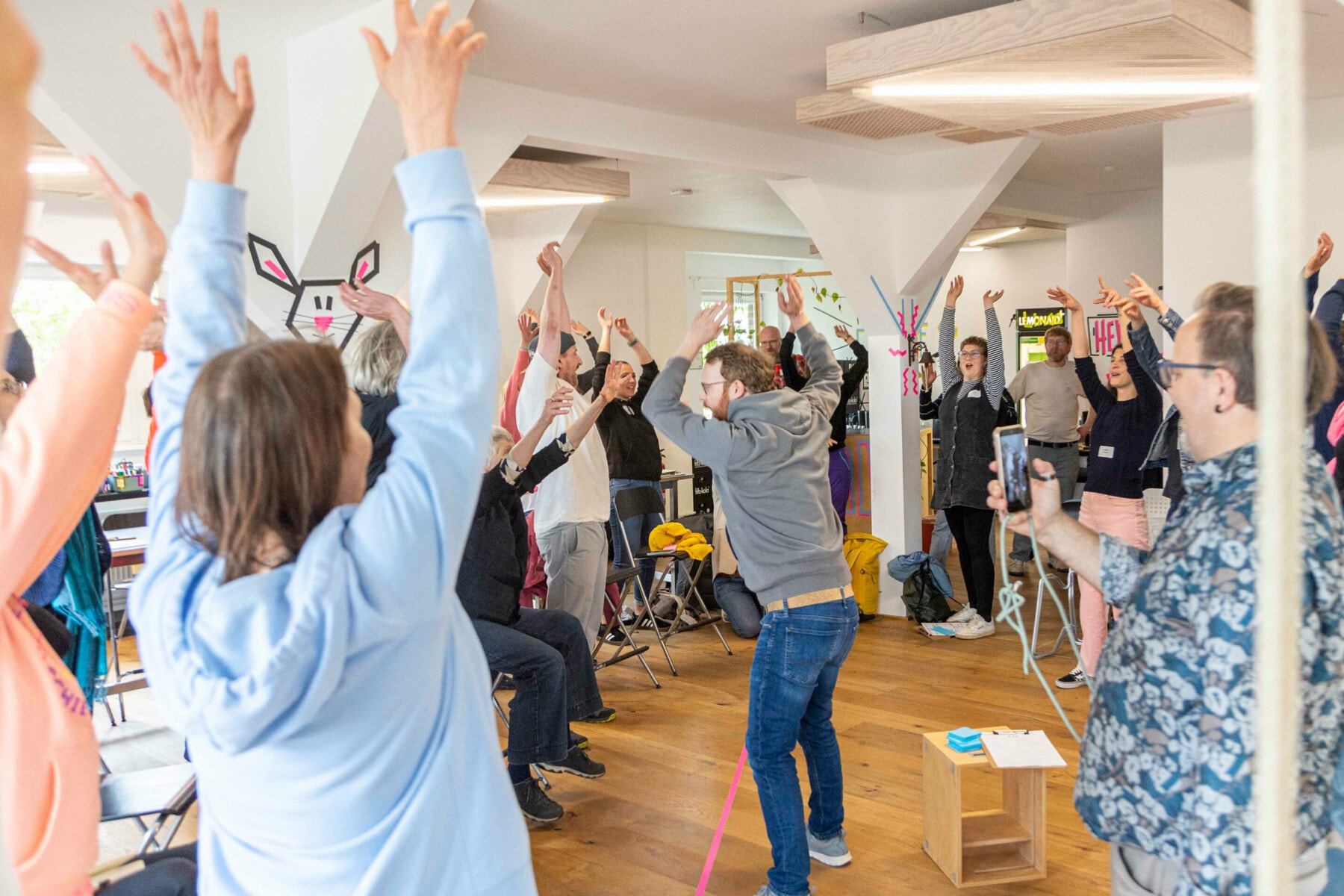 Teilnehmende eines Workshops stehen im Kreis. Ein Facilitator in der Mitte animiert die Gruppe, die Arme zu heben. Eine Person im rechten Bildrand filmt die Szene mit dem Smartphone.