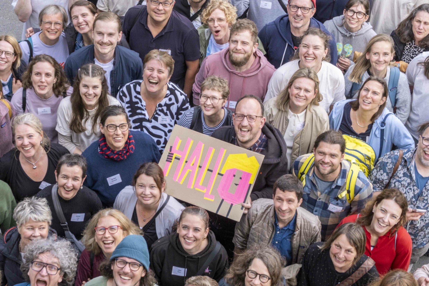 Große Gruppe von Menschen, die in die Kamera lächeln, eine Person hält ein Schild mit der Aufschrift 'HALLO' in bunten Buchstaben