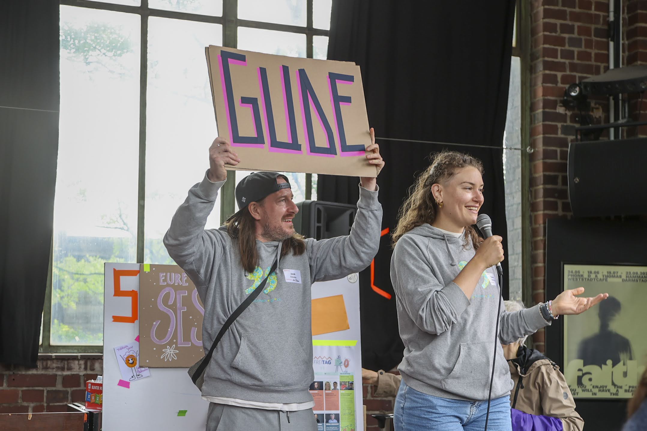 Zwei Personen bei der Eröffnung der KOON25 Konferenz, ein Mann hält ein Schild hoch, eine Frau spricht in ein Mikrofon, beide tragen graue Pullover mit buntem Aufdruck