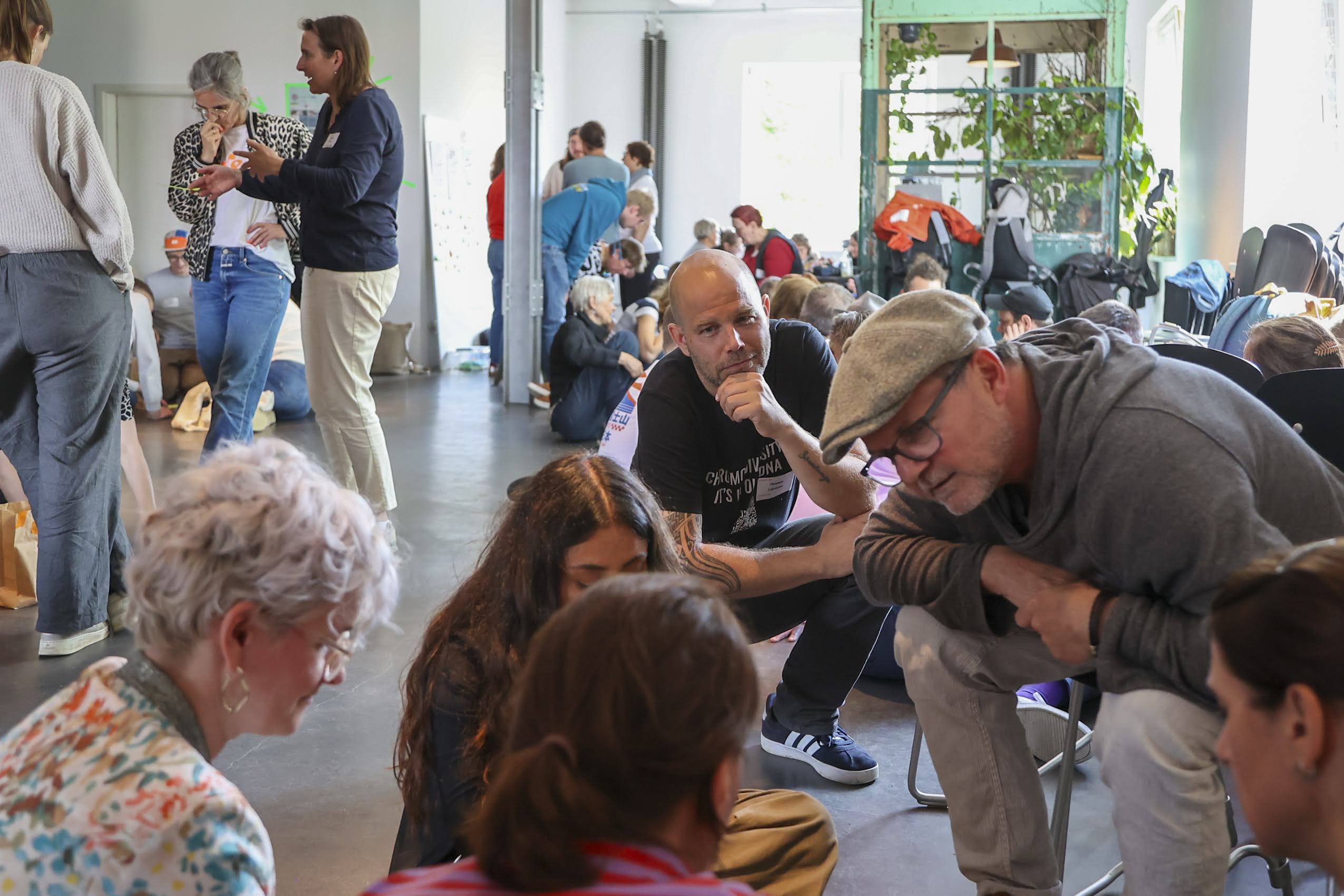 Teilnehmer eines Workshops auf der KOON25 diskutieren in kleinen Gruppen in einem hellen, offenen Raum. Einzelne Gruppen sitzen auf dem Boden, andere stehen. Im Mittelpunkt des Bildes hockt ein männlicher Facilitator und schaut einer Gruppe interessiert zu.