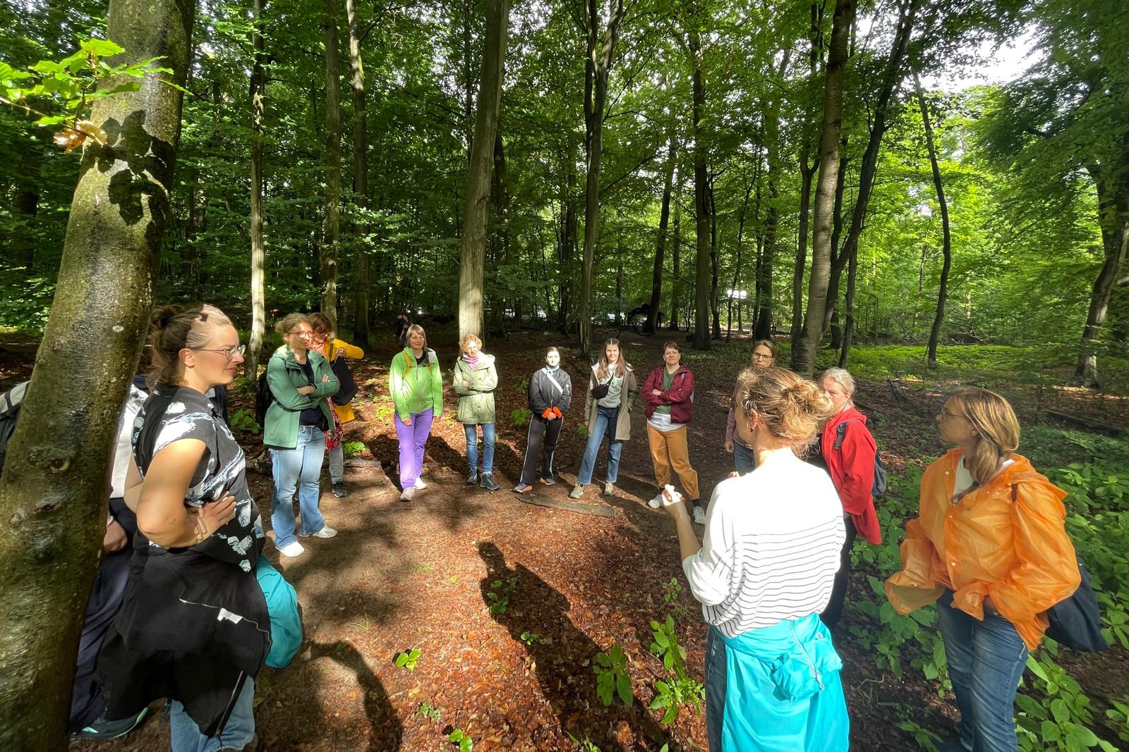 Teilnehmerinnen des Outdoor-Facilitation-Workshops der KOON25 stehen im Wald in einem Kreis und hören einer Frau zu, die spricht