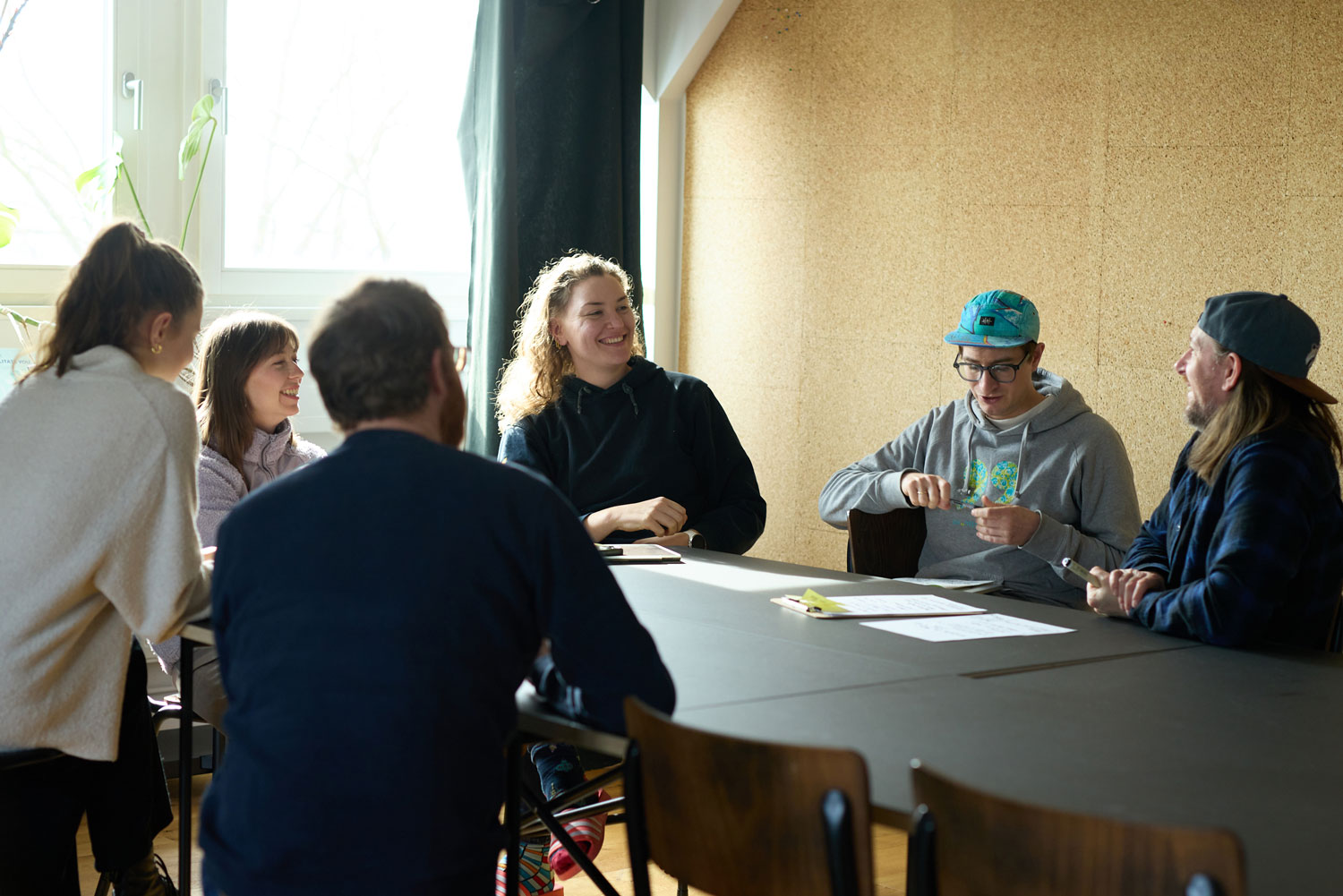 Gruppe von Menschen sitzt vor einer Korkwand um einen Tisch und unterhält sich. Einzelne Personen reden und lachen.